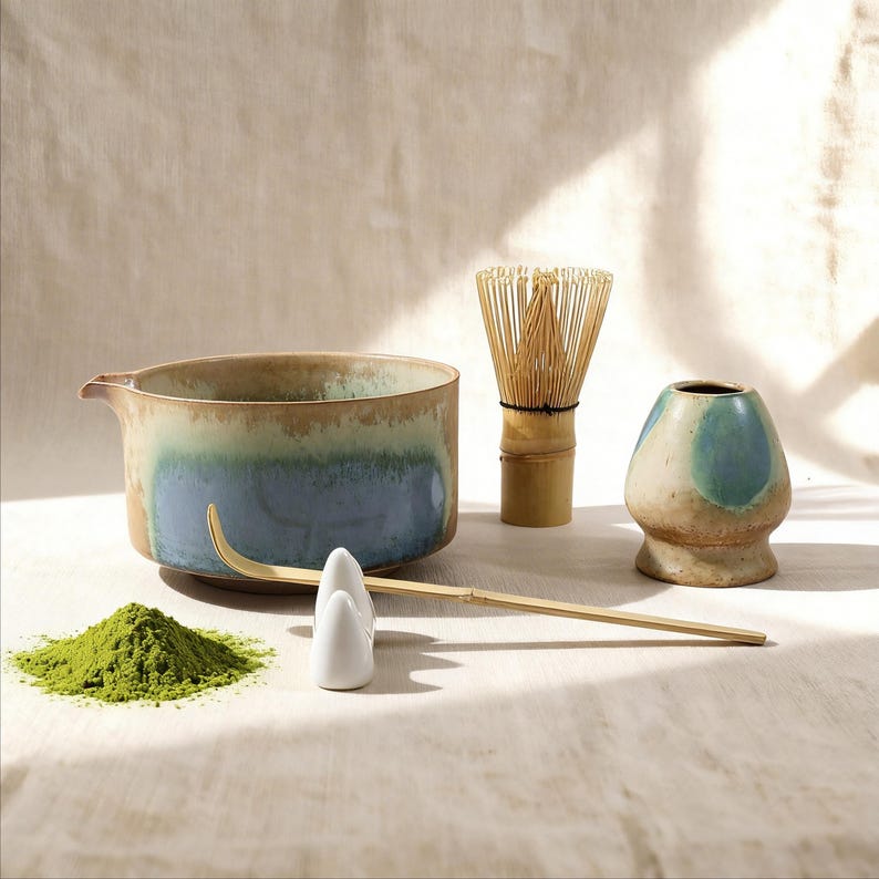 May include: A matcha tea set featuring a ceramic bowl with blue and brown glaze, a bamboo whisk, a whisk holder, a scoop, and a small ceramic container. A pile of green matcha powder is also present.