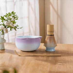 May include: A matcha tea set featuring a lavender and white ceramic bowl, a bamboo whisk, and a bamboo scoop. The bowl has a scalloped rim. The whisk and scoop are made of natural bamboo.