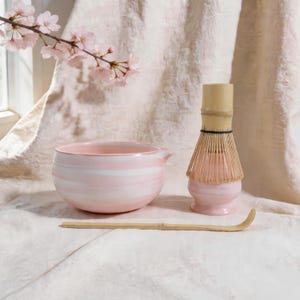 May include: A pink and white ceramic matcha bowl, bamboo whisk, and scoop are arranged on a light-colored surface. The bowl has a spout and a swirled design. The whisk is made of bamboo. A branch of pink cherry blossoms is in the background.