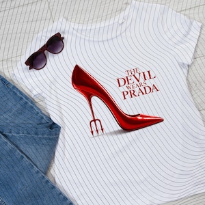 May include: White t-shirt with a red high heel shoe design and the text "The Devil Wears Prada". The shoe has a trident heel. A pair of sunglasses rests on the shirt. Blue jeans are in the image.