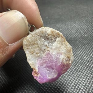 Pode incluir: Um pingente bruto e não polido com uma pedra mineral natural com uma divisão de cores distinta. A parte superior é uma estrutura cristalina opaca branca e bege, enquanto a parte inferior é um cristal rosa vibrante e translúcido.
