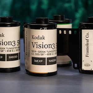 May include: A collection of Kodak Vision3 500T film rolls. The film canisters are white with black tops and feature the Kodak logo and product information. The image includes a strip of exposed film.