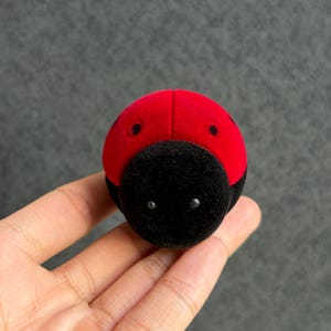 Può includere: Un piccolo portagioie arrotondato a forma di coccinella con una parte superiore rosso vivace dalla consistenza vellutata, caratterizzata da due punti neri e una sezione frontale nera tinta unita vellutata con due piccoli accenti circolari chiari.