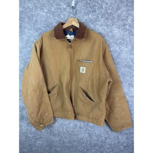 May include: A tan work jacket with a brown corduroy collar, a full zipper, and multiple pockets. The jacket features a small square logo on the chest and is hanging on a wooden hanger.