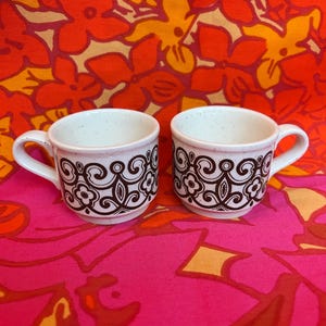 May include: Two white ceramic coffee cups with brown floral designs. The cups have small handles and a speckled texture. The background is a vibrant floral pattern in shades of red, orange, and pink.