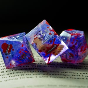 Puede incluir: Tres dados poliédricos con un efecto veteado azul y morado, salpicados de rojo. Los dados tienen números plateados y un diseño único en el interior. Los dados están sobre un libro.