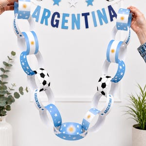 Puede incluir: Guirnalda festiva de cadena de papel con el diseño de la bandera argentina y balones de fútbol. La guirnalda se sostiene sobre un fondo blanco, con una pancarta que dice "ARGENTINA" en letras azules.