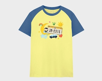 May include: A yellow short-sleeve t-shirt with blue raglan sleeves. The center features a graphic with a license plate reading JM-2315, a crescent moon, a sun, a small blue van, a rainbow, a heart, and a cartoon character illustration.