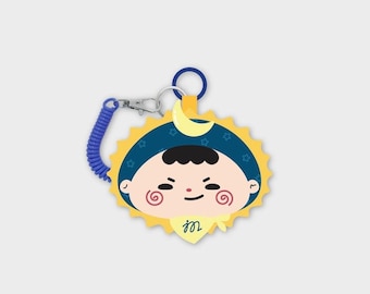 May include: A circular keychain featuring a cartoon character with a dark blue hood, a yellow sun-shaped border, and a yellow crescent moon. The character has pink spiral cheeks and a yellow neckerchief with the letter m. Includes a blue coil cord.