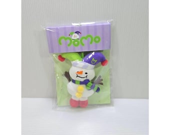 May include: A plush snowman toy in clear plastic packaging with a purple header card featuring the text momo. The snowman wears a green and purple jester hat, a striped scarf, and has an orange carrot nose, two yellow buttons, and pink feet.