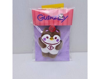 May include: A small brown and white plush penguin toy wearing a white jersey with the letters JG in maroon. The penguin has purple cheek spots, a yellow beak, and a small red basketball on its head. It is packaged in a clear plastic bag with a pink and purple header card featuring the text GUINZLY.