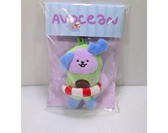 May include: A plush keychain featuring a purple and green character shaped like an avocado, wearing a red and white striped life preserver. The item is sealed in clear plastic packaging with a header card displaying the text Avocean.