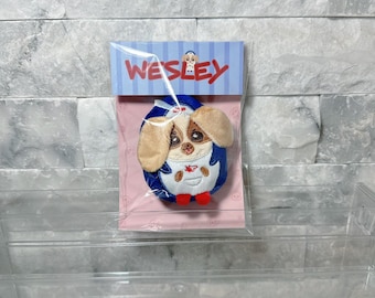 May include: A small plush toy shaped like a dog with tan ears, large brown eyes, and a white belly, wearing a blue and white outfit with red feet. The toy is packaged in a clear plastic bag with a header card featuring the text WESLEY.