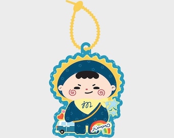 May include: A decorative acrylic charm featuring a cartoon character wearing a dark blue hooded outfit with a yellow scarf. The character has red spiral cheeks and is positioned next to a small blue van and a rainbow with the text JUMMO.