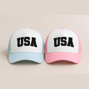 Two baseball caps with the word "USA" printed on them, one in blue and one in pink.