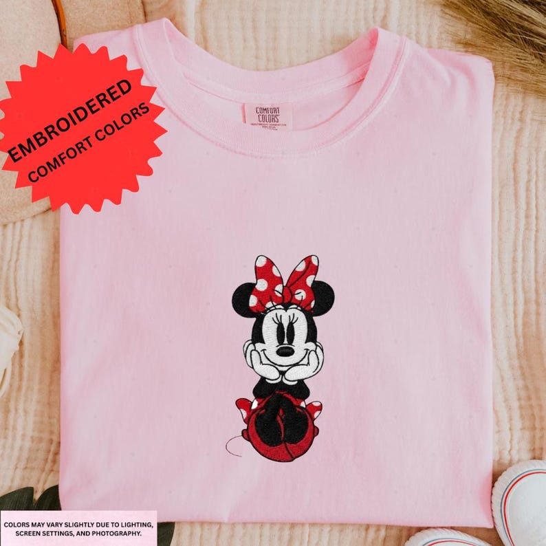 A pink t-shirt with a Minnie Mouse embroidered design is displayed on a surface.
