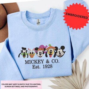 A light blue t-shirt with a graphic design featuring the faces of Mickey Mouse and Goofy, along with the text "MICKEY & CO. Est. 1928".