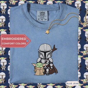 a blue t-shirt with a graphic of two characters, a baby Yoda and a young alien, along with a star-shaped charm. The t-shirt is displayed against a background with a repeating pattern of the same characters.