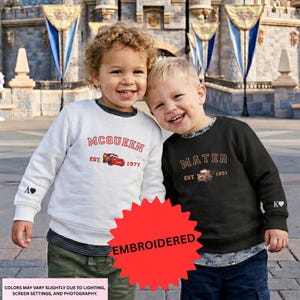 Two young children, a boy and a girl, are standing together and smiling in front of a castle. They are wearing sweatshirts with the text "McQueen" and "Mater" printed on them.