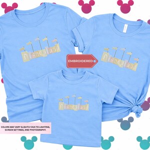 three blue t-shirts with the word "Disneyland" printed on them, along with a red tag that says "EMBRODERED". The t-shirts appear to be family-sized, suggesting they are intended for a family or group of people. The background of the image is a white surface with colorful Mickey Mouse ears scattered across it.