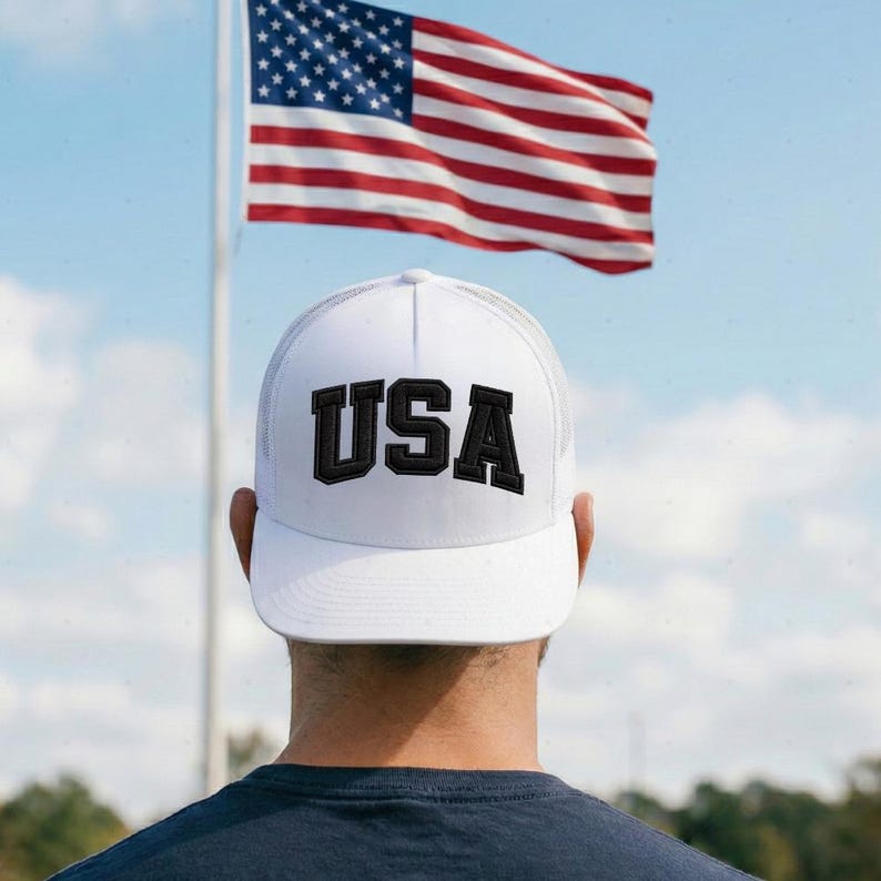 A person wearing a white baseball cap with the word "USA" printed on it, standing in front of an American flag.