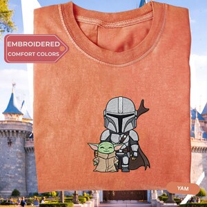 a red t-shirt with a graphic of a character and a child, likely representing the Mandalorian family from the Star Wars franchise.