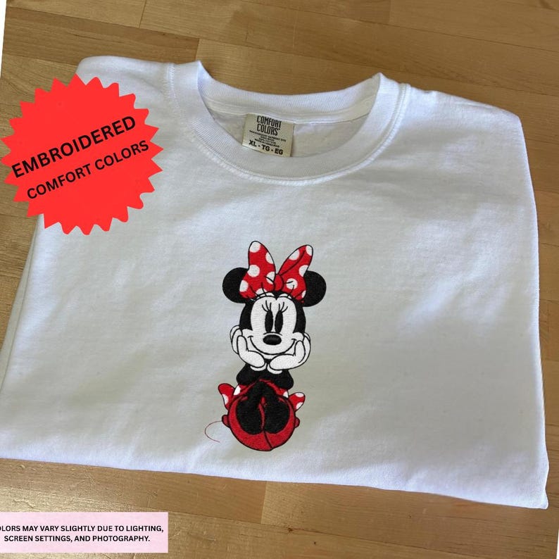 A white t-shirt with a Minnie Mouse design printed on it, along with a red starburst label indicating that the colors are embroidered.