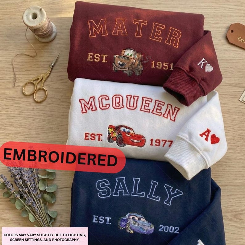 three embroidered sweatshirts with the character Sally from the animated movie Cars. The sweatshirts are arranged on a wooden surface, with a spool of thread and a pair of scissors nearby. The text on the sweatshirts reads "EMBRODERED" and "EST. 2002", indicating that they are custom-made and have been embroidered. The colors of the sweatshirts vary slightly due to lighting, screen settings, and photography, and the text suggests that the colors may not be consistent across all images.