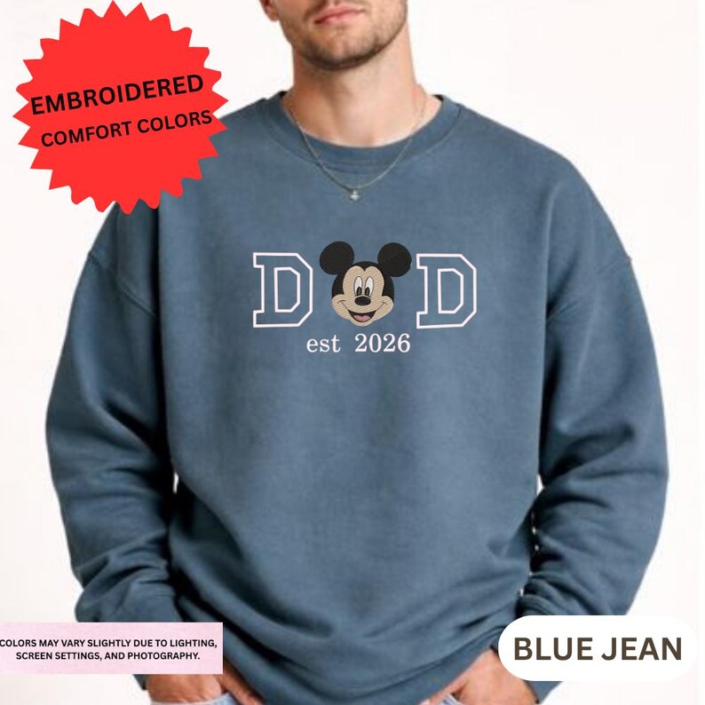 A man wearing a blue sweatshirt with a Mickey Mouse logo and text that says "DAD est 2026".