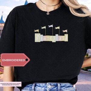 A person wearing a black t-shirt with the word "Disneyland" embroidered on it, standing in front of a Disney castle.