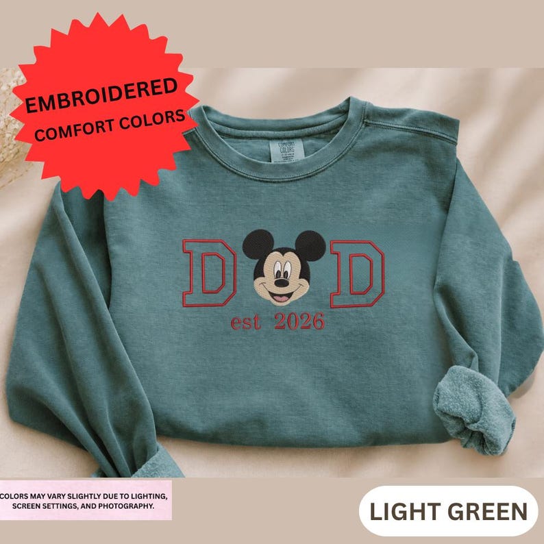 a green long-sleeved shirt with a red Mickey Mouse logo and text that reads "DAD est 2026".