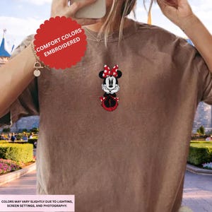 A person wearing a brown t-shirt with a Minnie Mouse embroidered on it, standing in front of a Disney-themed background.