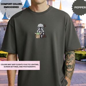 A person wearing a gray t-shirt with a graphic of a character from the Star Wars franchise, specifically the Mandalorian, standing in front of a castle-like structure.