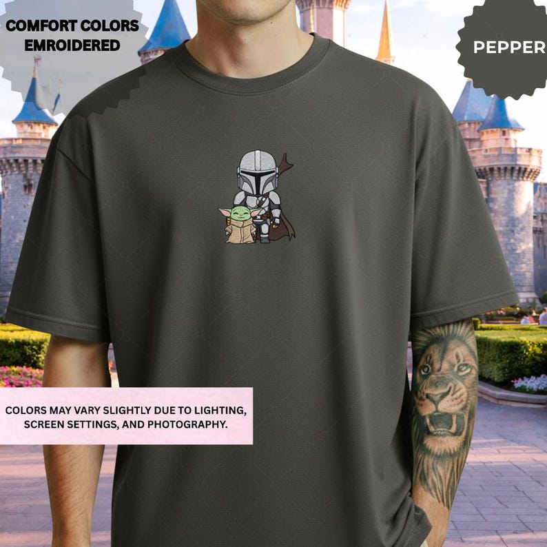 A person wearing a gray t-shirt with a graphic of a character from the Star Wars franchise, specifically the Mandalorian, standing in front of a castle-like structure.