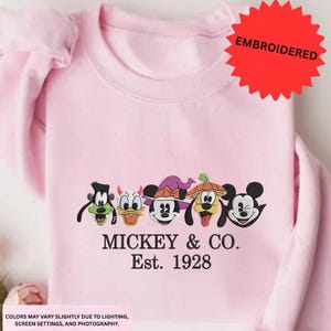 A pink t-shirt with a graphic design featuring the characters Mickey Mouse, Donald Duck, Goofy, and Pluto, along with the text "MICKEY & CO. Est. 1928".