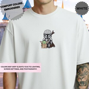 A white t-shirt with a graphic of a character and a smaller character, likely representing a character from a popular media franchise. The t-shirt is described as being embroidered and features a castle in the background.