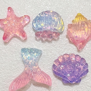 Peut inclure: Cinq breloques en résine irisée comprenant une étoile de mer rose, une méduse bleue et violette, un coquillage rose et jaune, une queue de sirène bleue et rose, et une coquille Saint-Jacques violette. Chaque pièce a une finition brillante et pailletée.