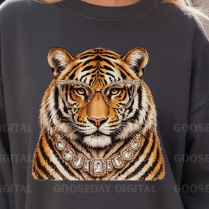 Könnte beinhalten: Ein anthrazitfarbenes Sweatshirt mit Rundhalsausschnitt und einem zentralen Tiger-Motiv, das eine Katze-Augen-Brille mit goldenem Rahmen sowie eine große, verzierte goldene Halskette mit klaren, rechteckigen Steinen trägt. Der Text GOOSEDAY DIGITAL ist wiederholt.
