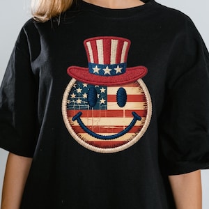 May include: Black t-shirt featuring a smiley face design with an American flag theme. The smiley face wears a red, white, and blue Uncle Sam hat. The design includes the American flag and a stitched border.