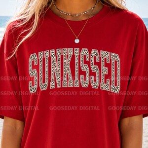 以下が含まれることがあります： 大きなアーチ状の大学風フォントで「SUNKISSED」という文字がプリントされた赤い半袖Tシャツ。文字はベージュと茶色のヒョウ柄で、無地の赤い生地に対して細い白い輪郭線が施されています。