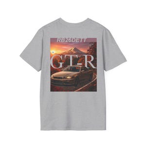 Pode incluir: Camiseta cinza com um gráfico de um carro em uma estrada sinuosa com um fundo de montanha. O texto "RB26DETT" está acima da imagem, e "GTR" está escrito em letras grandes sobre o carro.