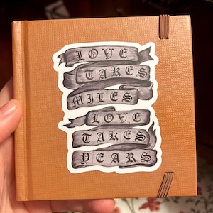 May include: A small, square, orange-brown journal with a matching ribbon bookmark. A sticker on the cover reads "LOVE TAKES MILES LOVE TAKES YEARS" in a gothic font on gray banners. The journal is held in a hand.