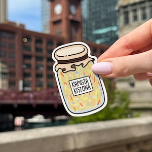 May include: A die-cut sticker featuring a glass jar filled with shredded yellow and orange vegetables, topped with a tan lid and cloth cover. A white rectangular label on the front of the jar displays the black text KAPUSTA KISZONA.