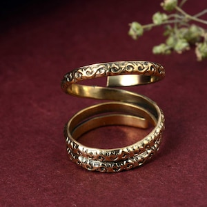 May include: A gold-colored spiral ring with an intricate, repeating pattern. The ring wraps around the finger in a unique design, showcasing detailed craftsmanship. The ring is displayed against a deep red background.
