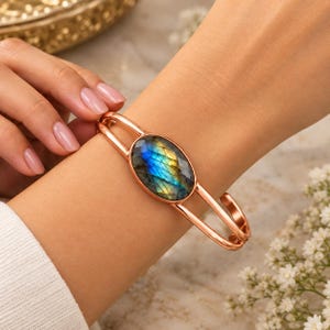 May include: A rose gold-toned cuff bracelet featuring a central oval-shaped labradorite gemstone. The stone displays iridescent blue and yellow flashes within its dark, faceted surface. The bracelet band consists of two parallel metal wires.
