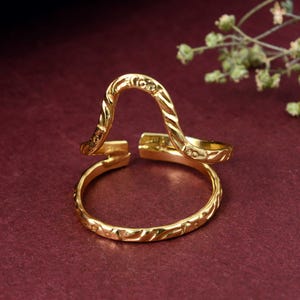 May include: A gold-colored adjustable ring with an open design. The ring features an arched top and a band with etched details. The ring is displayed on a burgundy surface, with a sprig of greenery in the background.