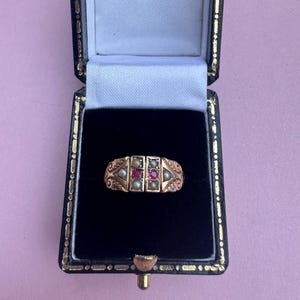 May include: A gold ring featuring two central red gemstones flanked by four small white pearls, set within a decorative metal band with engraved scrollwork details, presented inside a black velvet-lined jewellery box with a white interior lid.