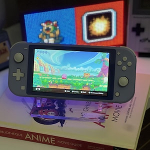 May include: A light blue handheld gaming console featuring a screen displaying a colorful landscape with a pink character. The device has a directional pad, two analog sticks, and four action buttons. It is positioned on a clear acrylic stand.