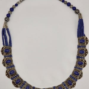 May include: A silver-toned necklace with oval, deep blue gemstones and matching blue beads. The necklace has a decorative, ornate design with a hook clasp. The necklace is a statement piece.