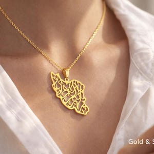 Puede incluir: Un collar con colgante dorado que presenta un mapa estilizado de un país con intrincada caligrafía. El collar cuelga de una delicada cadena sobre una camisa blanca. Las palabras "Gold & Silver" son visibles en la imagen.
